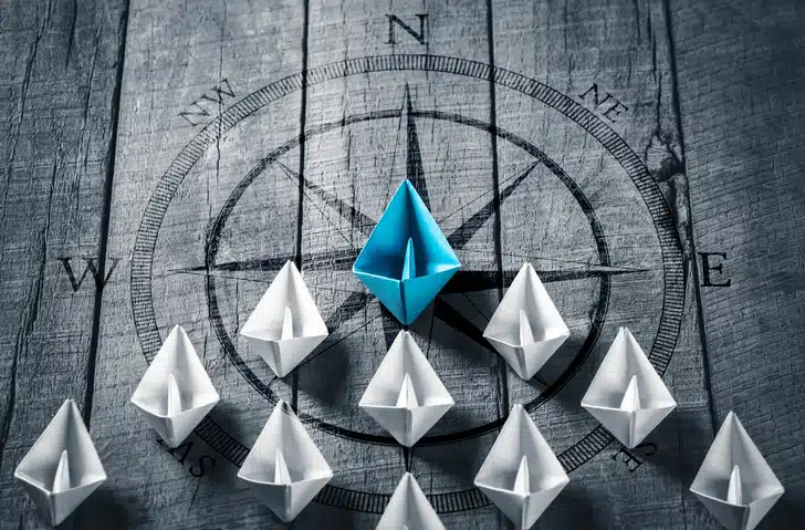 Photo of seven white paper boats being led by a blue paper boat. Represents talent strategy and talent optimization, which can help an organization develop their leaders.