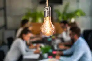 Team working together in background, with antique light bulb in foreground. Fresh ideas keep employees engaged, which is important to talent strategy.
